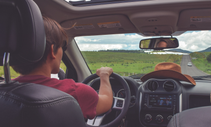 Person driving a car on a rural road, with a brown cowboy hat on the dashboard and an open sunroof above
