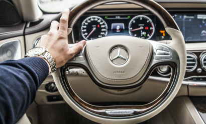 Person's left hand on the steering wheel of a Mercedes-Benz car, dashboard and instrument panel visible in the background.