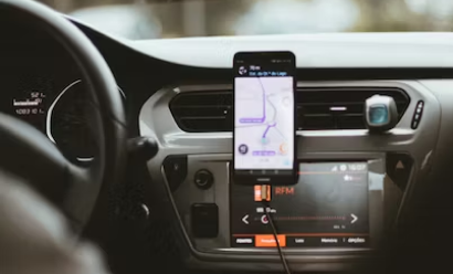 Smartphone mounted on a car dashboard displaying GPS navigation. The car's steering wheel and part of the center console with audio controls are also visible.